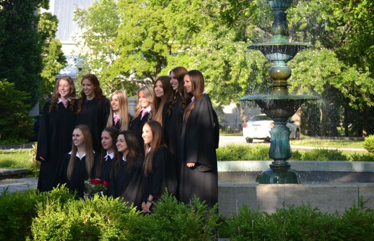 Groupe de jeunes femmes en toge de graduation, posant devant une fontaine, cérémonie extérieure, environnement verdoyant, célébration de réussite scolaire, Fondation Saint-Sacrament, Collège Saint-Sacrament, lumière naturelle, ambiance festive et élégante.